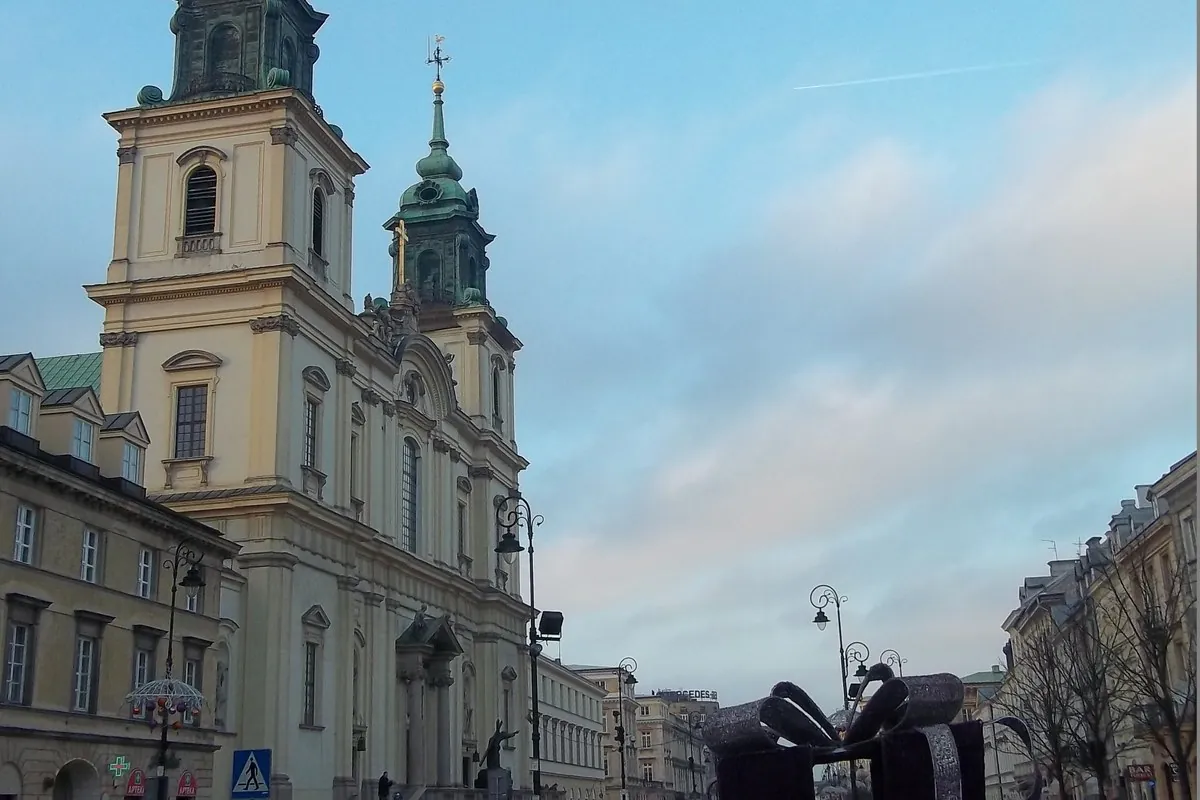 The twin-towered, cream-coloured front of Holy Cross Church seen from a Warsaw street at dusk with a green copper roof