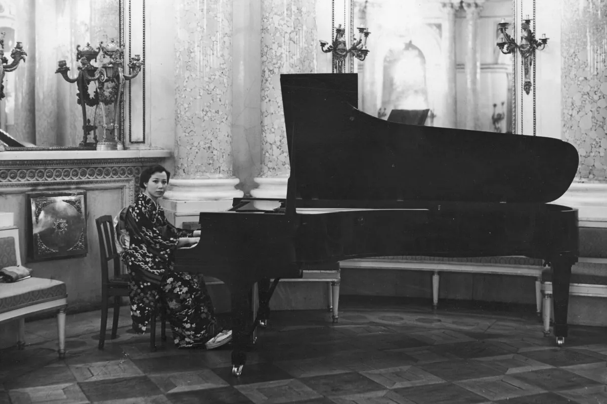 A black-and-white photograph of a woman in a kimono playing a grand piano in a columned hall with candelabra on the walls